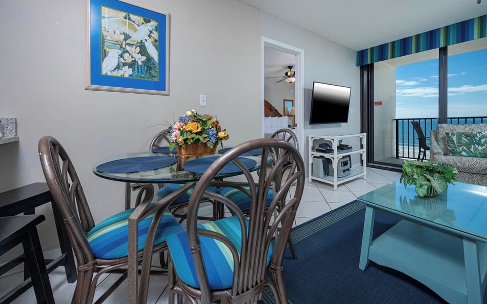 The living area features a glass dining table, colorful decor, and a TV, with a stunning beach view through the sliding doors