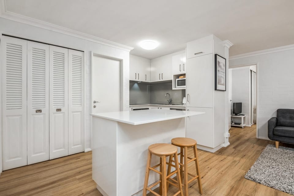 White kitchen island with two wooden stools contrasts with gray backsplash and light wood flooring.