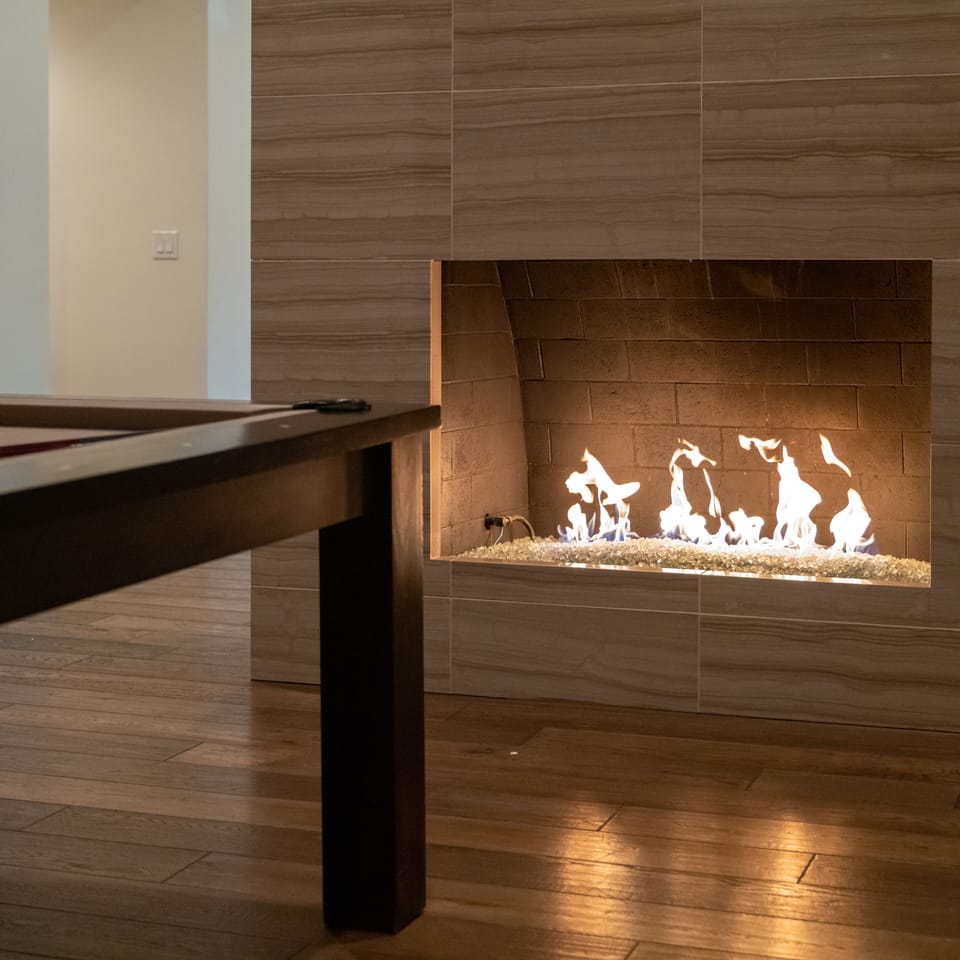 A flickering fireplace casts a warm glow next to the pool table, illuminating the natural wood floors and creating a cozy ambiance perfect for gathering and relaxation.
