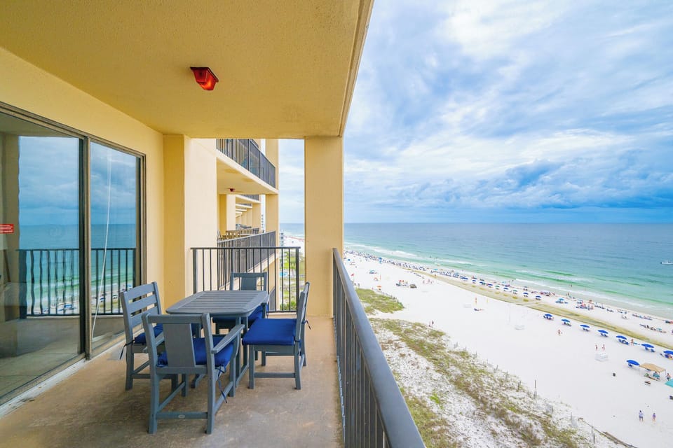 The balcony features a table with red and blue chairs, offering a direct view of the beach and turquoise waters below
