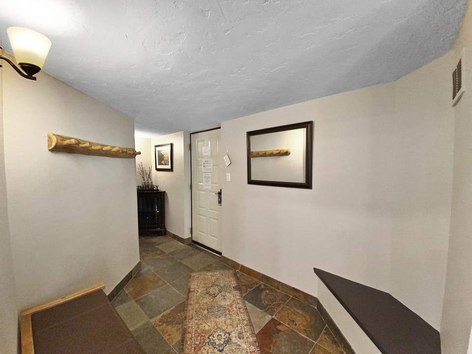 A small entryway featuring a bench, a rug, a wall-mounted coat rack, and a framed mirror. The door has several papers attached, and a side table with decor items is visible in the corner.