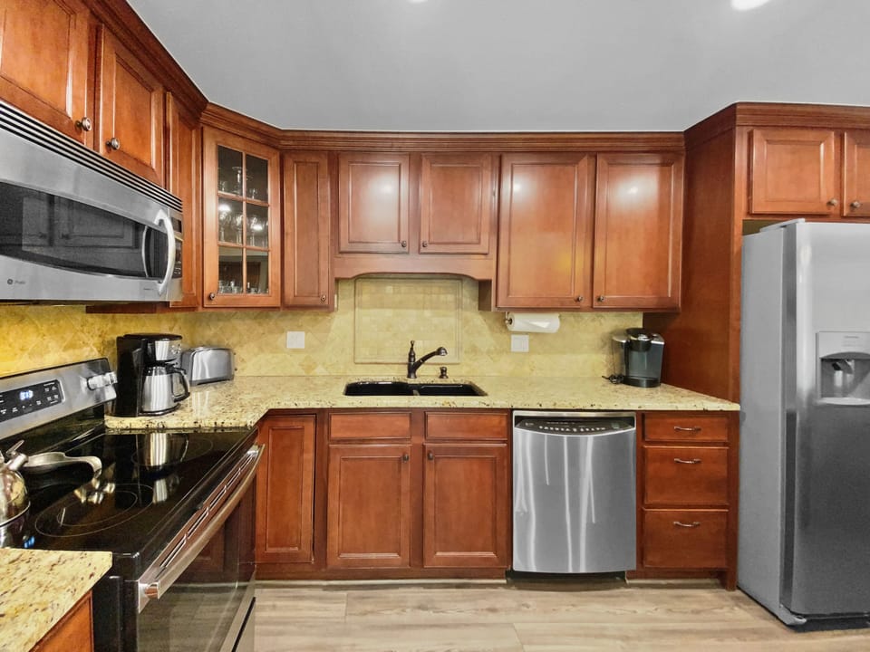 A modern kitchen with wooden cabinets, stainless steel appliances, granite countertops, a built-in microwave, dishwasher, sink, refrigerator, coffee maker, and kettle.