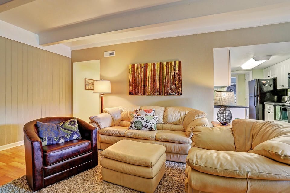 A living room with a leather armchair, a matching sofa, and an ottoman. The space features a forest-themed painting, a standing lamp, and an open view into the adjoining kitchen.