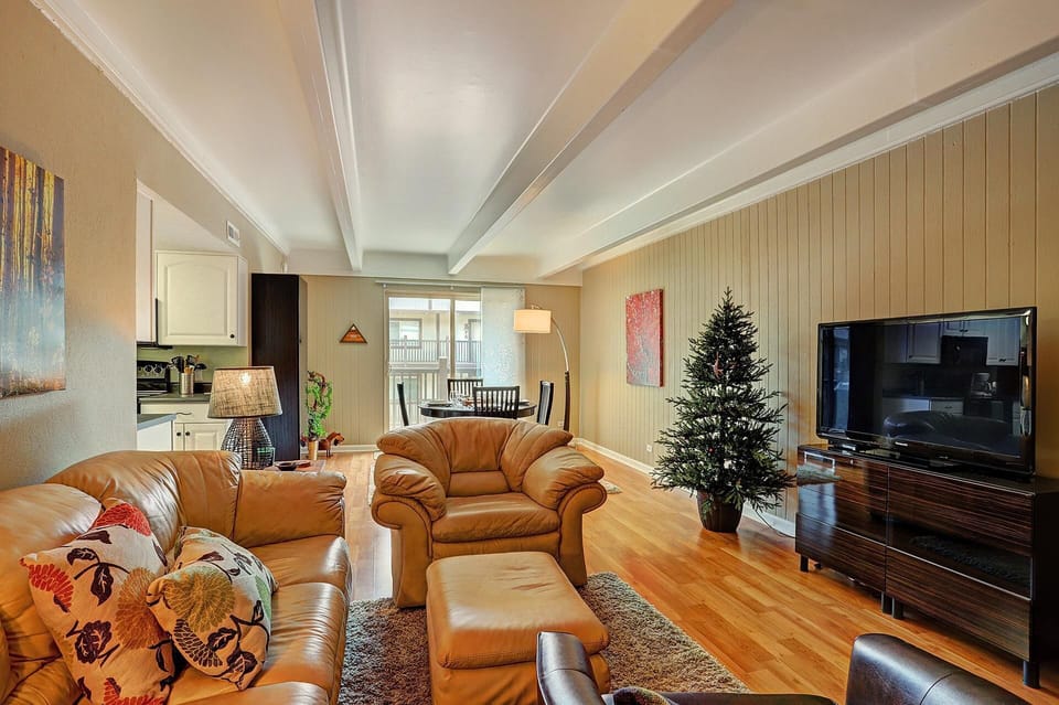 A living room with beige sofas, an armchair, a TV, a Christmas tree, and artwork on the walls. The space has wooden flooring and an open dining area in the background.