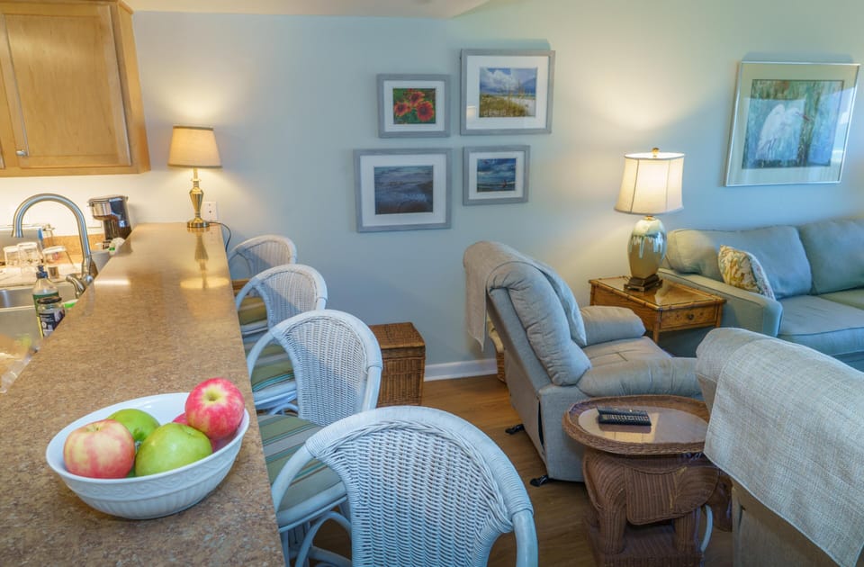 Granite countertop with white wicker bar stools overlooks living area featuring a blue-gray recliner and sofa.
