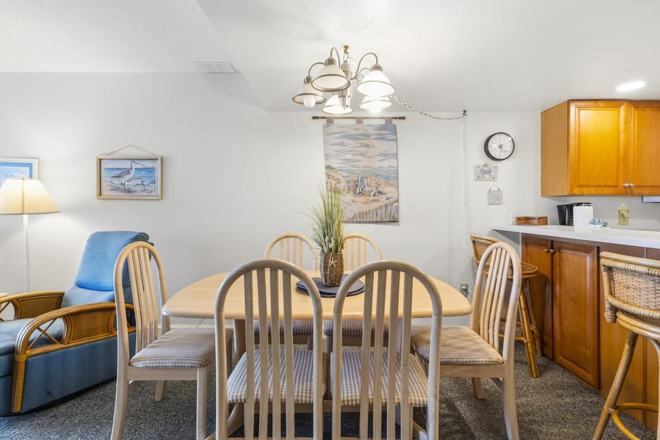 Dining area with light wood table and six padded chairs, rattan armchair, coastal wall art, and overhead light fixture.