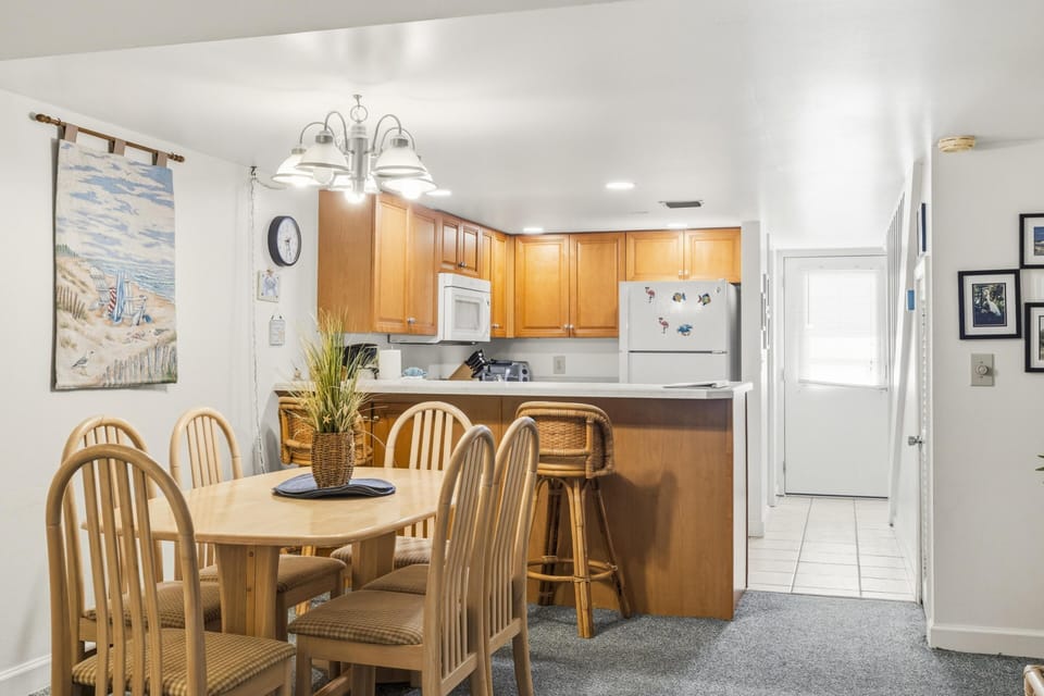 Dining area with light wood table and six chairs, adjacent to kitchen bar with wicker stools and oak cabinetry.