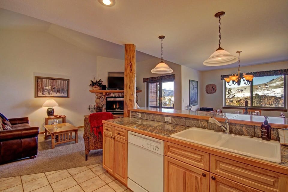 A cozy open-plan living room and kitchen area with wooden cabinetry, modern appliances, a leather sofa, a stone fireplace, large windows, and mountain views.