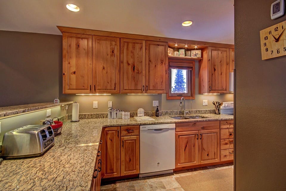 A kitchen with wooden cabinets, granite countertops, a dishwasher, a double-slice toaster, and a microwave. There are small decorative items above the window. A clock is mounted on the wall.