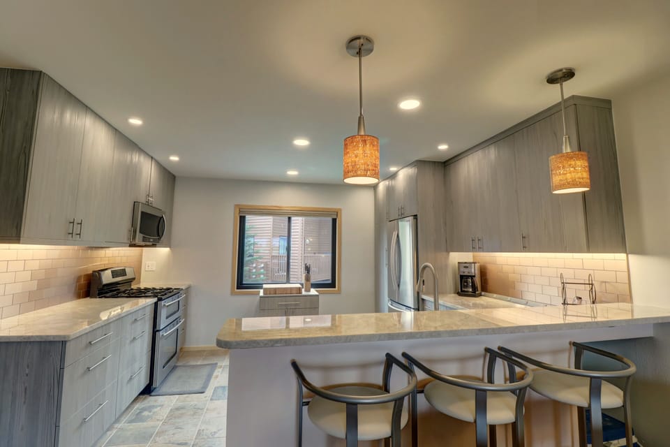 Modern kitchen with grey cabinets, stainless steel appliances, a central island with bar seating, pendant lights, and a window over the sink.