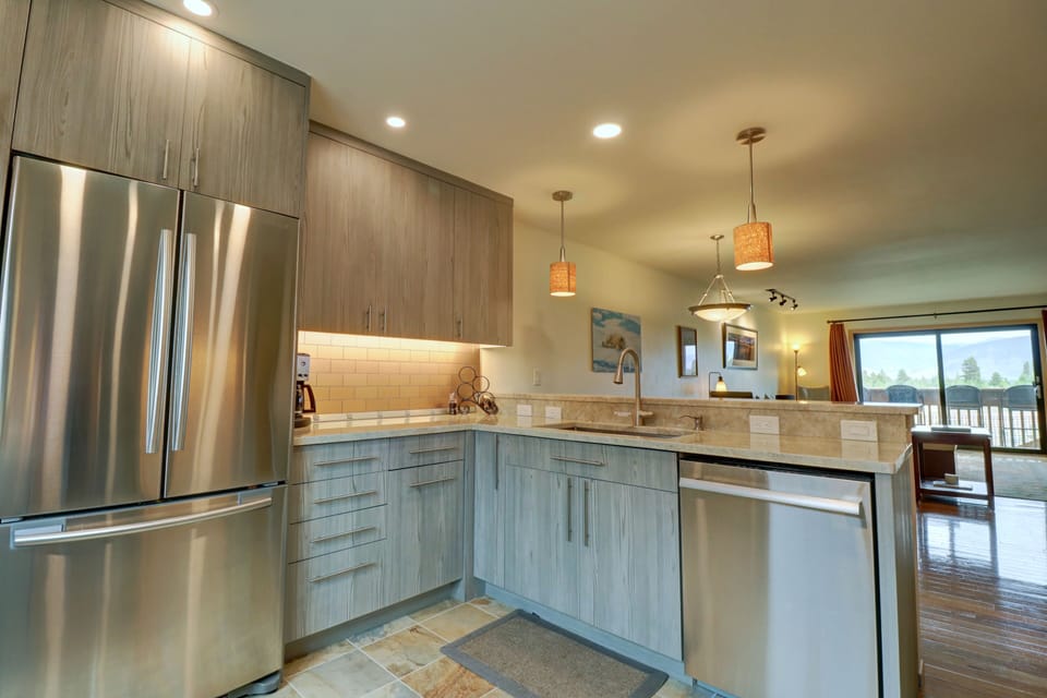 Modern kitchen with stainless steel appliances, gray wood cabinets, light granite countertops, pendant lights, and a view into a living area with sliding glass doors leading to a balcony.