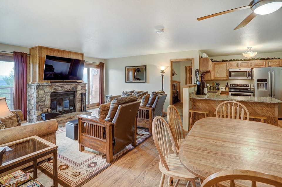 Open-concept living and dining area with wooden furniture, a stone fireplace, wall-mounted TV, and a kitchen in the background. Ceiling fan and hardwood floors are present.