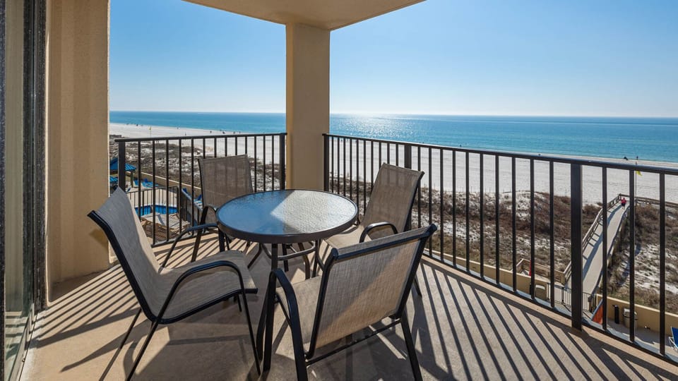A balcony features a round table and four chairs, offering a serene view of the beach and sparkling blue waters