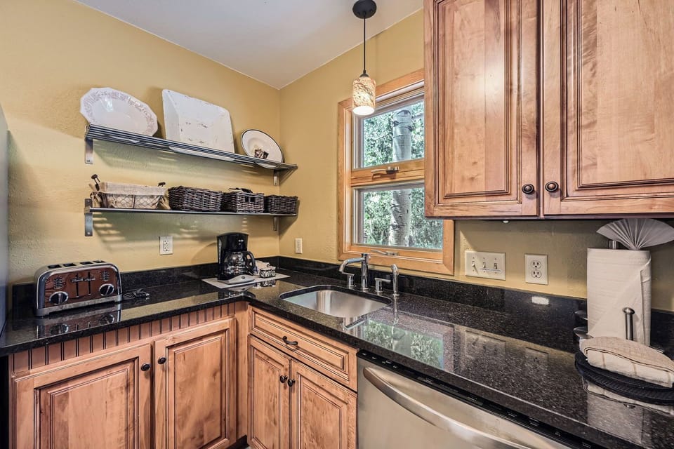 A small corner kitchen with wooden cabinets, a black granite countertop, a sink, a window, a toaster, a coffee maker, and a shelf holding plates and baskets.