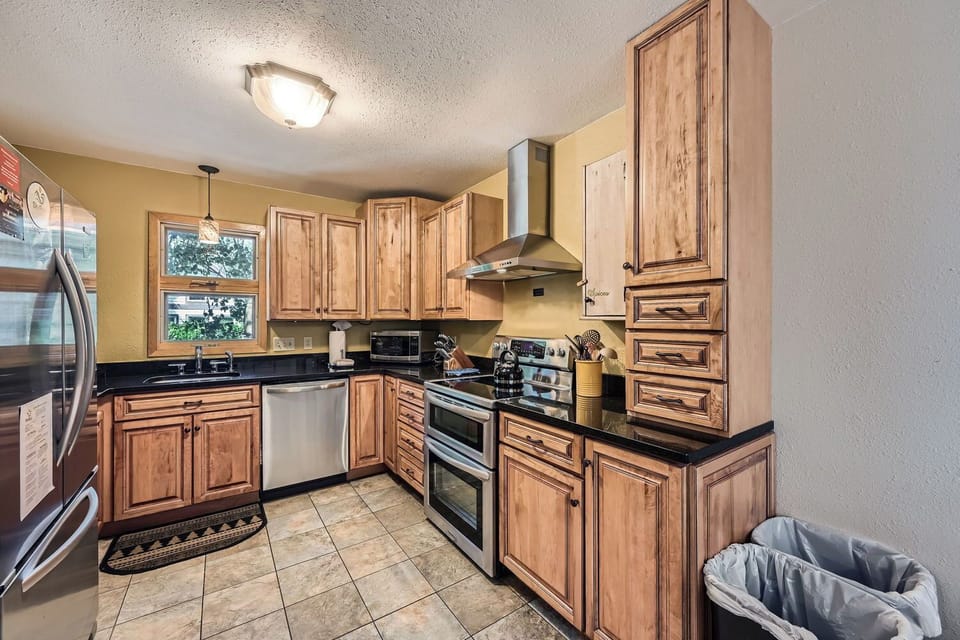 A kitchen with wooden cabinets, stainless steel appliances, including a refrigerator, stove, and dishwasher, a small window above the sink, and tiled floors. A trash bin is visible in the corner.