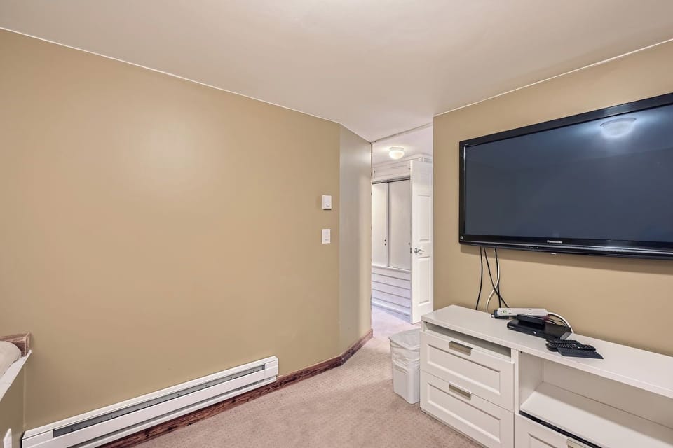 A room with beige walls features a flat-screen TV mounted on the wall, a white dresser with drawers, and a small trash bin. A door in the background leads to another area.