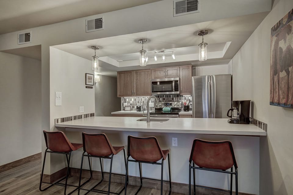 A modern kitchen with a central island, four bar stools, stainless steel appliances, and wooden cabinetry. The ceiling has recessed lighting and pendant lights hang above the island.