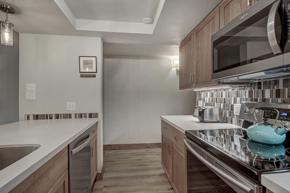 Modern kitchen with light wooden cabinetry, stainless steel appliances, a teal kettle on the stove, and tiled backsplash. Walls include minimalistic decor and a pendant light hangs above the countertop.