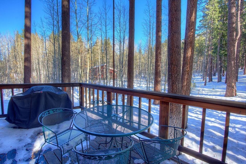 Snow-season deck with outdoor table seating, covered grill, railing, trees, and neighboring building.