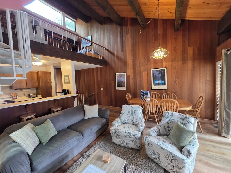 Open living area with sofa, armchairs, dining table, breakfast bar, kitchen cabinets, loft railing, ceiling fan, and hanging light.