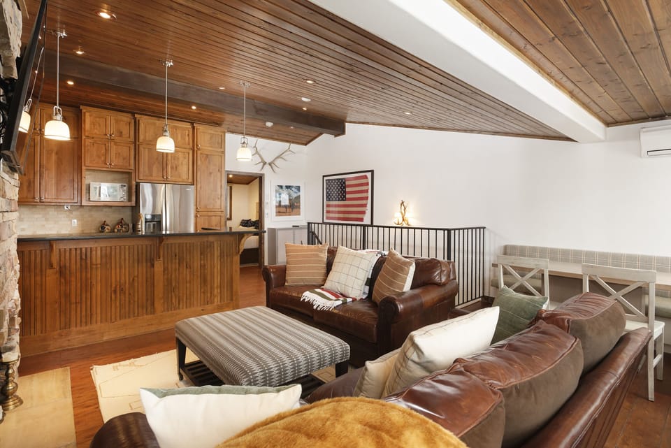 A cozy living room with a brown leather sofa, checkered ottoman, and wooden ceiling. The background shows a kitchen area with wooden cabinets and a bar counter. An American flag decorates the wall.