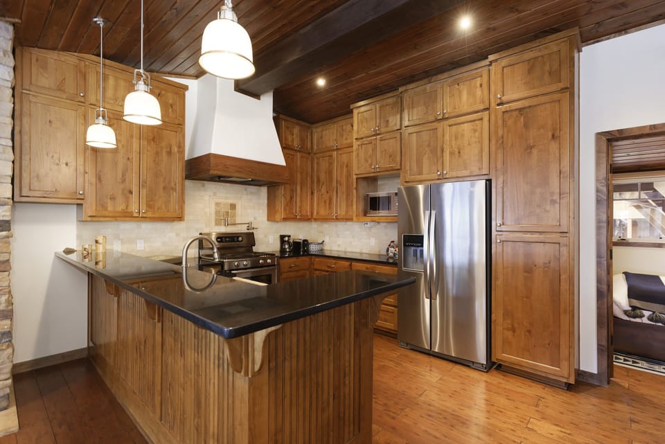 A modern kitchen with wooden cabinets, stainless steel appliances, a black countertop, and three pendant lights hanging over the island. The ceiling is wooden with recessed lighting.