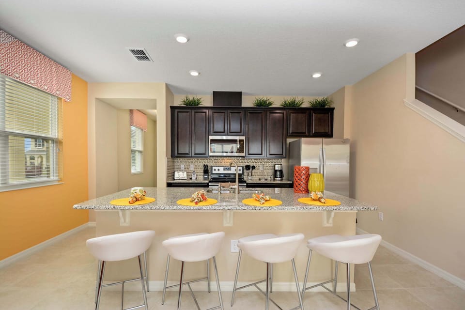 Sunlit kitchen nook with an orange accent wall, granite counters, and four white bar stools for a sunny, casual vibe.