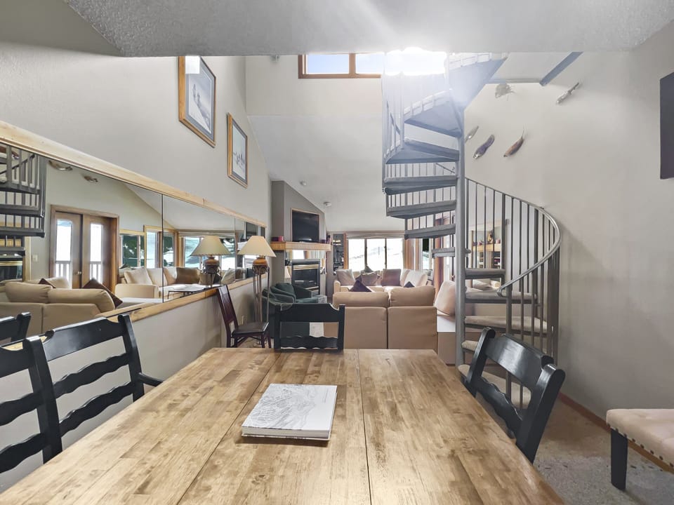 A living space features a spiral staircase, wooden dining table with chairs, and a living room with beige sofas. The room has large mirrors and wall-mounted decorations. Natural light filters in from windows.