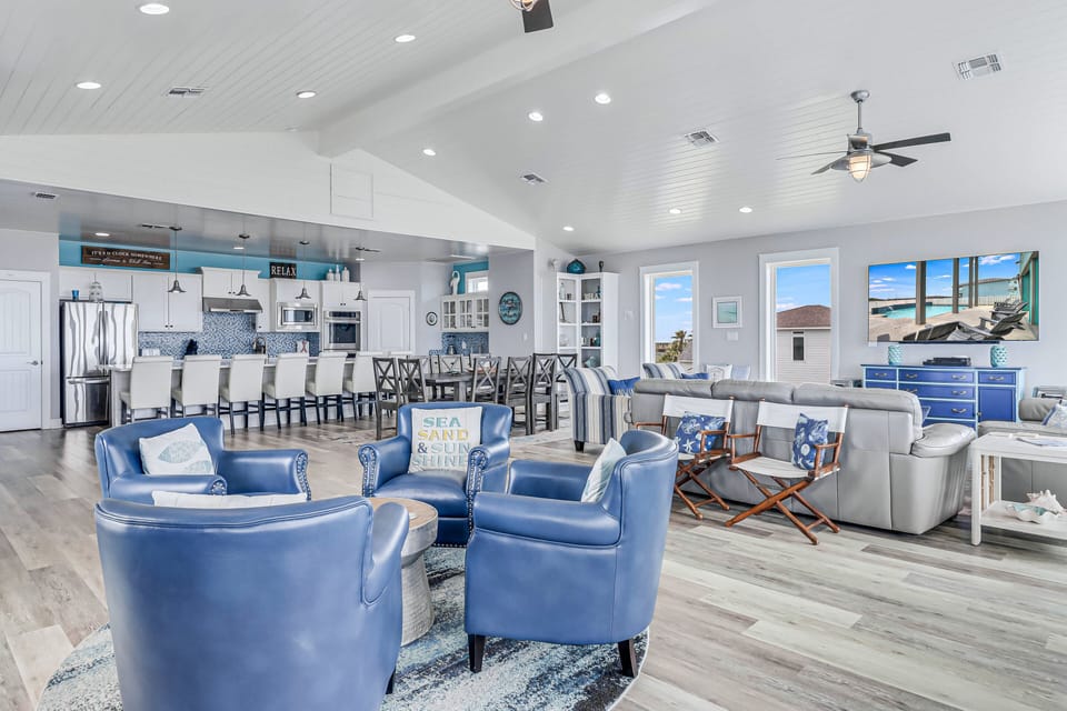 Your expansive open-concept living space features coastal-inspired blue furnishings, gleaming kitchen, and stunning ocean views through floor-to-ceiling windows.