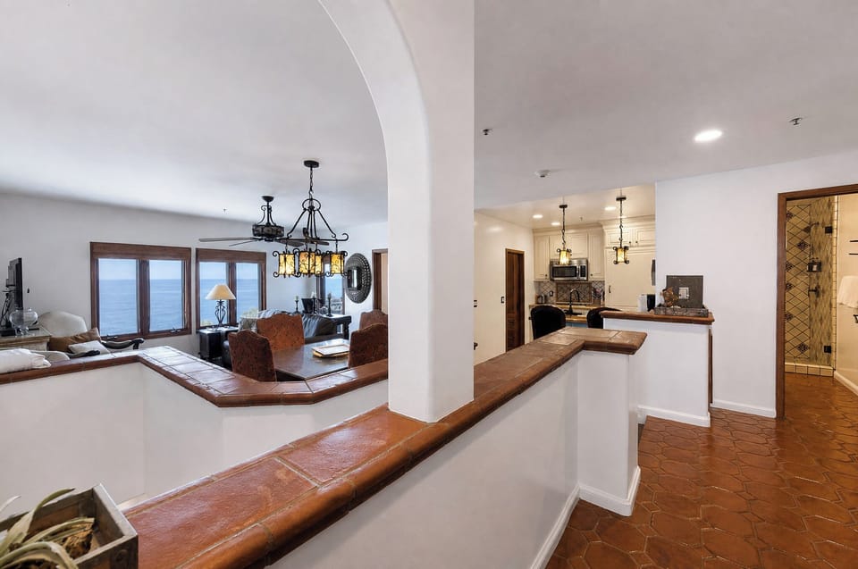 Admire ocean views through elegant arched windows as you gather in your open-concept living space, where warm wood tones, decorative pendant lighting, and terracotta tile floors create a welcoming Mediterranean atmosphere.