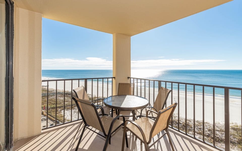 The balcony features a glass-top table surrounded by four chairs, offering a panoramic view of the tranquil beach and water