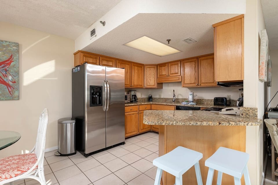 The kitchen features warm wooden cabinetry, granite countertops, coffee maker, and a breakfast bar with seating for two