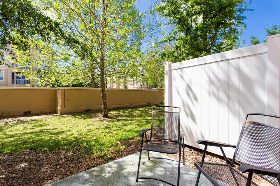 Two black metal mesh chairs rest on a shaded concrete slab beside a white privacy fence and green lawn.