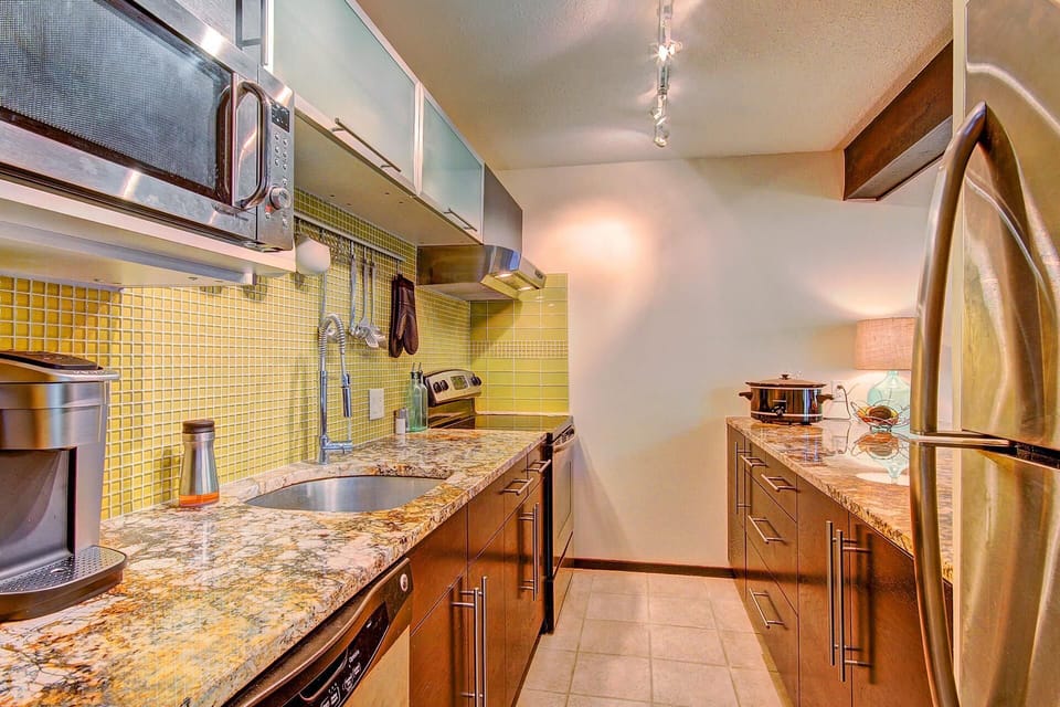 Modern kitchen with granite countertops, stainless steel appliances, yellow tile backsplash, under-cabinet lighting, and wooden cabinetry. Coffee machine on counter, and various kitchen utensils hung up.