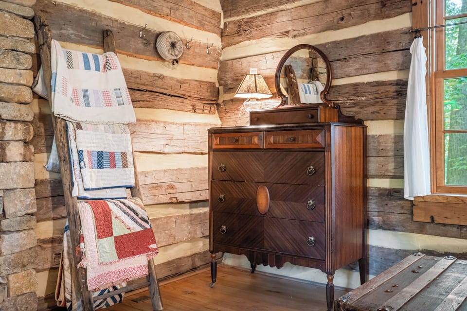 Charming log cabin room with vintage quilts, antique dresser, and woodland views—filled with warm details and timeless, nostalgic touches.
