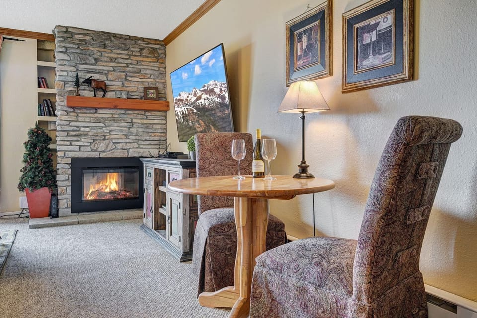 Small round wooden table set for two with patterned upholstered chairs beside a stone fireplace and wall-mounted TV.