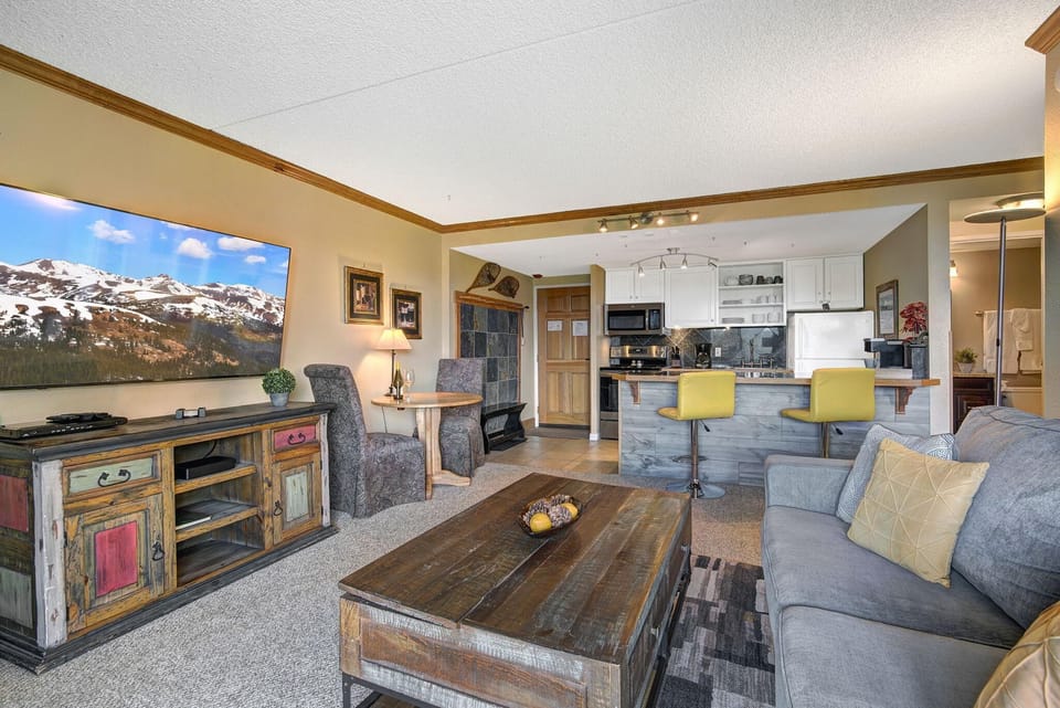 Living room with distressed wood media cabinet, gray sofa with yellow pillow, and ski-themed decor near kitchen bar.