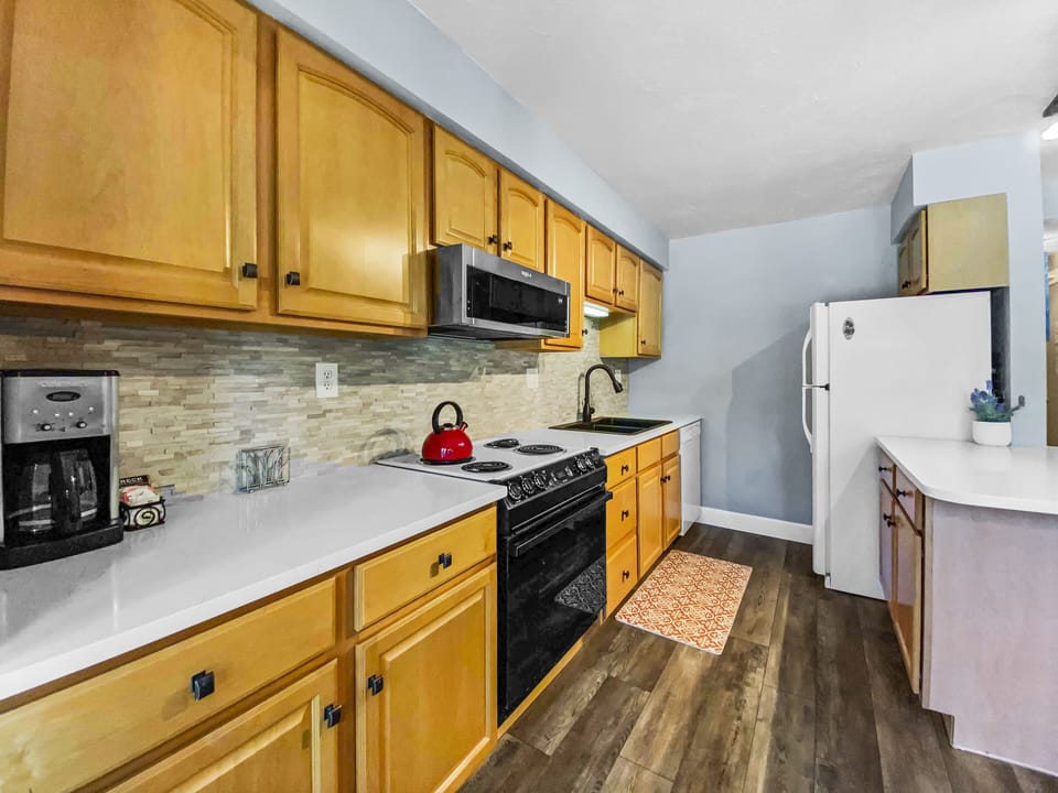 A modern kitchen with wooden cabinets, a white countertop, a black stove, a stainless steel microwave, a refrigerator, a coffee maker, a red kettle, and a patterned rug on the floor.