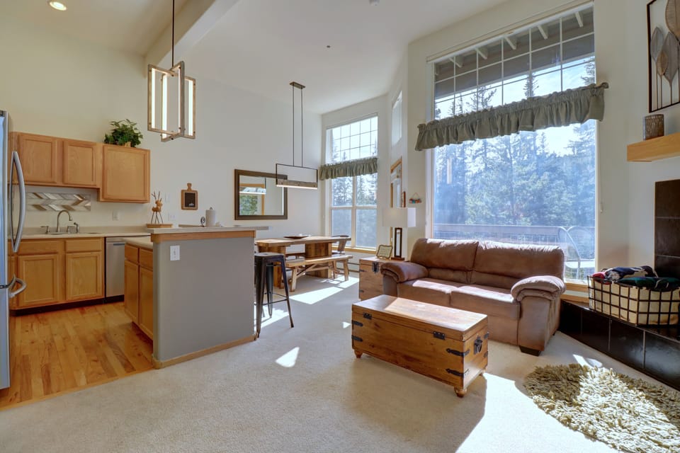 A bright, open-plan living room and kitchen area features wood cabinetry, a dining table, and a leather sofa near large windows with forest views. A wooden chest and shelves add a rustic touch.