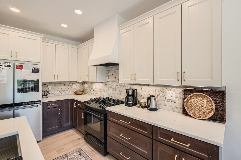 Modern kitchen with white upper cabinets, dark lower cabinets, a white range hood, brick backsplash, and stainless steel appliances. A coffee maker and wicker tray are on the countertop.