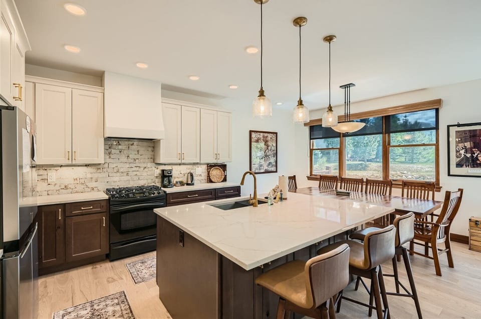 Modern kitchen with a large island and barstools, featuring white cabinets, stainless steel appliances, pendant lights, and an adjacent dining area with a wooden table and chairs.