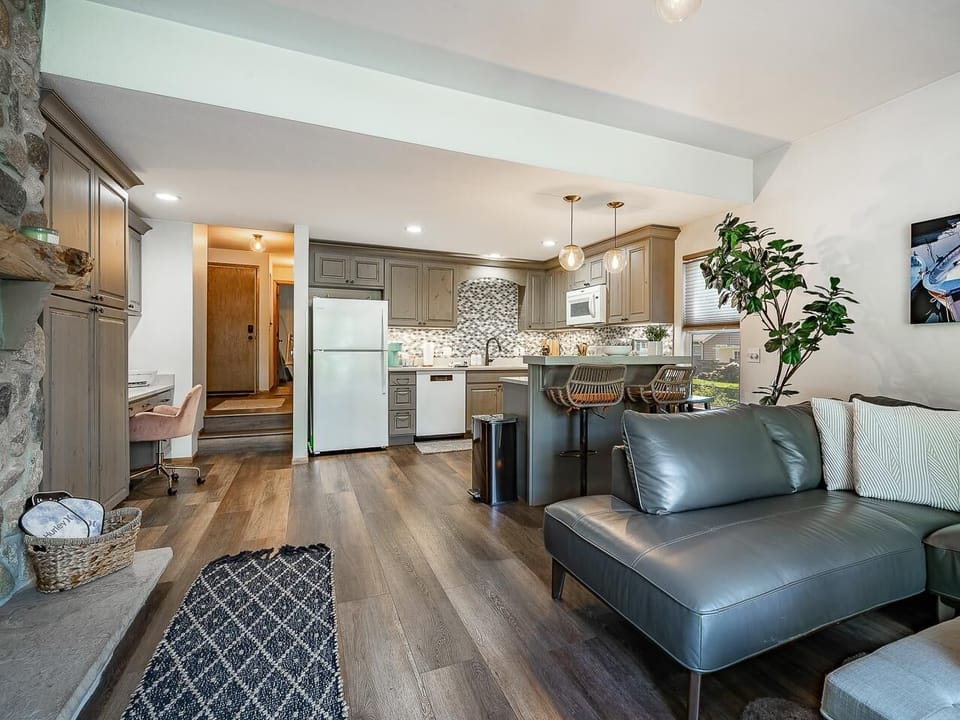 Open-concept living area with gray leather sectional, wood floors, stone fireplace, and kitchen island with wicker bar stools.