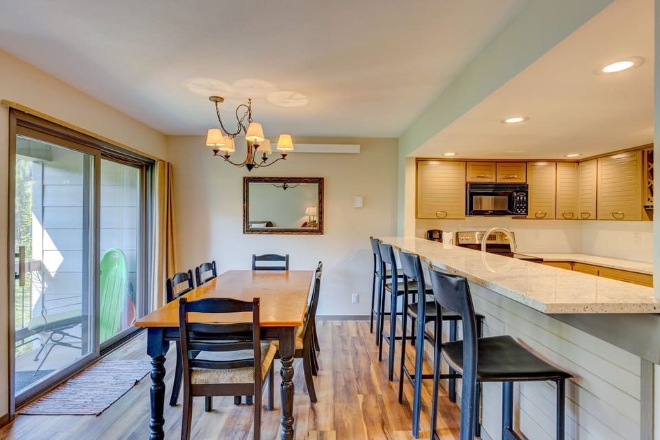 A dining area with a wooden table and six chairs, next to a kitchen with a breakfast bar and four bar stools. There's a chandelier above the table, a large mirror on the wall, and sliding glass doors on the left.