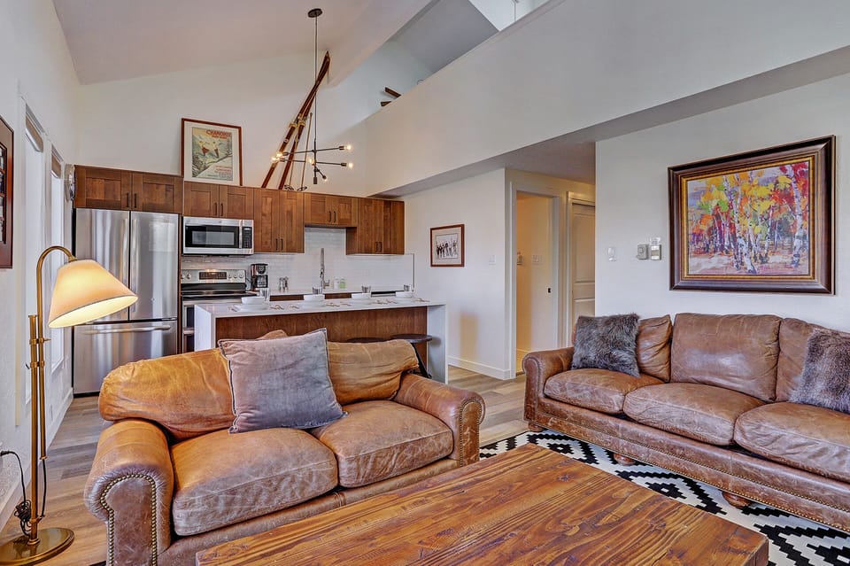 A cozy living room with brown leather couches, a wooden coffee table, patterned rug, modern kitchen in the background, a staircase leading to an upper level, and colorful wall art.