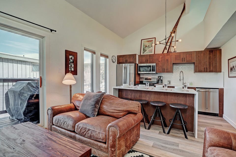 A cozy open-plan living area featuring a brown leather sofa, a kitchen with wooden cabinets, pendant lighting, and a breakfast bar with three stools. A sliding glass door leads to a small balcony with a grill.