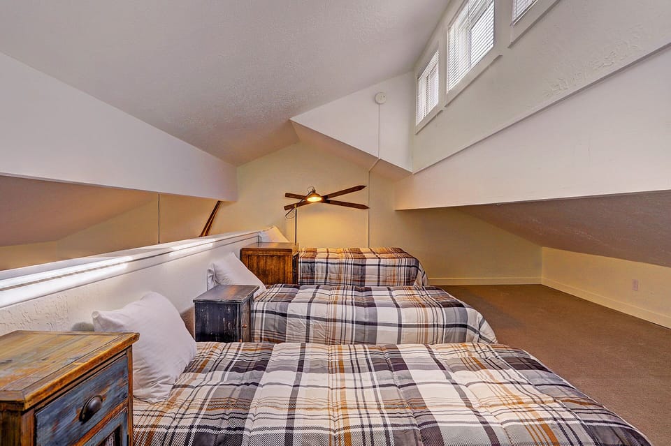An attic bedroom with two plaid-patterned beds, wooden nightstands, and a ceiling fan. The room has a sloped ceiling and windows on the upper wall.
