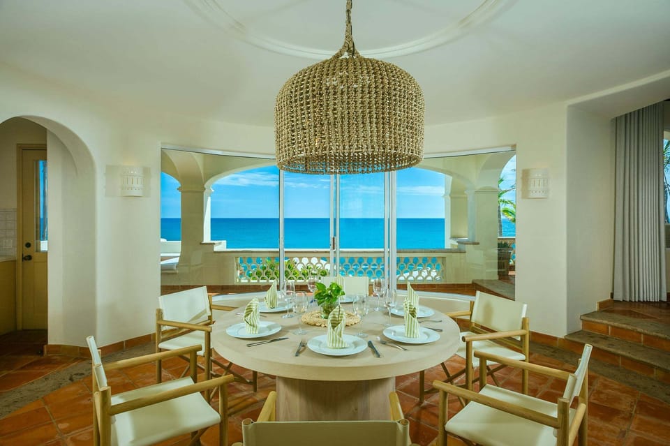 Indoor dining room with an elegant round table, wicker lamp, and large windows overlooking the sea.