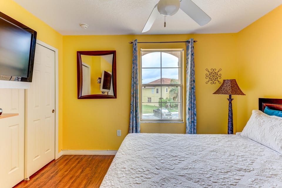 Bedroom with bright yellow walls, white quilted bedding, wood floor, window with blue patterned curtains, and ceiling fan.