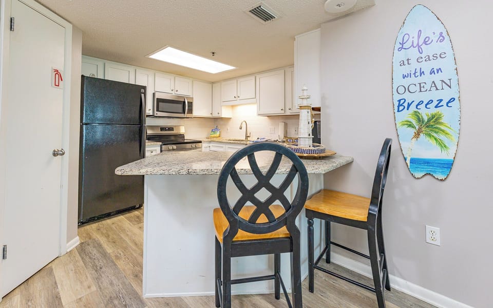 Kitchen features a breakfast bar with seating, granite countertops, and a vibrant surfboard sign adding coastal charm