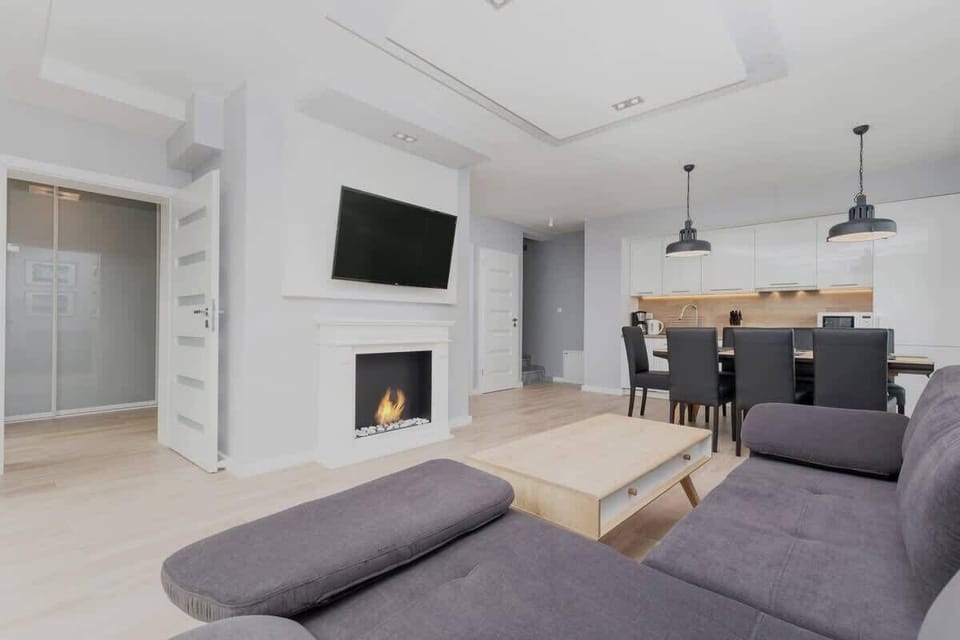 A spacious living room with a cozy sectional sofa, a wooden coffee table, a large wall clock, and abundant natural light coming through the sliding glass doors.